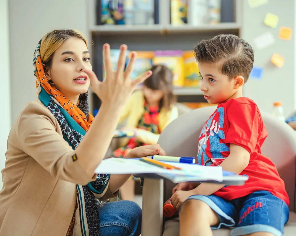 An educator in a headscarf counts on her fingers with a young boy during a one-to-one learning session