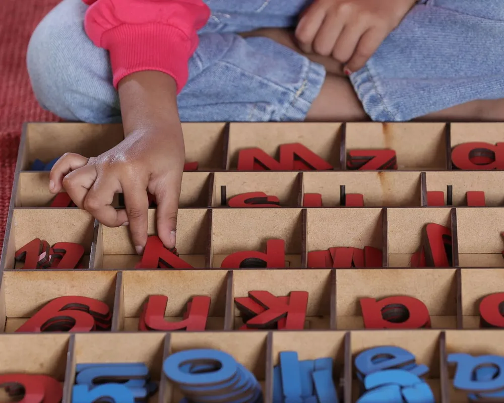 A child's hand picks wooden letters from a sorting tray during a fine-motor activity